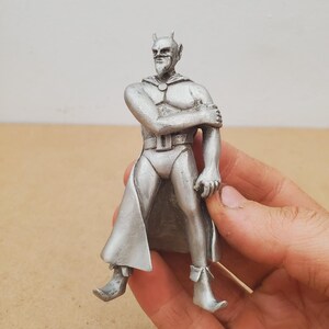 May include: A silver figurine of a devil-like character with horns, a cape, and boots. The figure is posed with one arm across its chest. The figurine is held in a hand.
