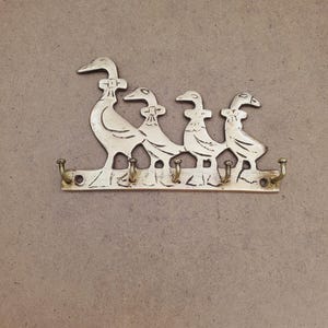 Puede incluir: Un llavero de pared de color latón con un diseño decorativo de cuatro gansos en fila. Cada ganso tiene un contorno detallado y un pequeño gancho debajo para colgar llaves. El llavero está montado sobre un fondo de tono neutro.