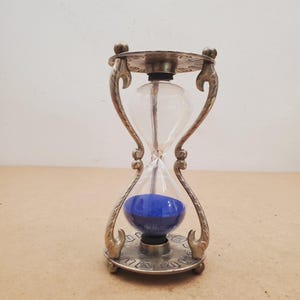 May include: An hourglass featuring a metallic frame with ornate, curved supports and circular top and bottom plates. The clear glass bulb contains vibrant blue sand, which is currently settled in the bottom section of the hourglass timer.