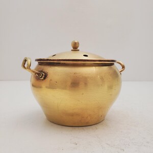 May include: A small, gold-colored brass pot with a lid and two handles. The pot has a round body and a slightly domed lid with small holes.