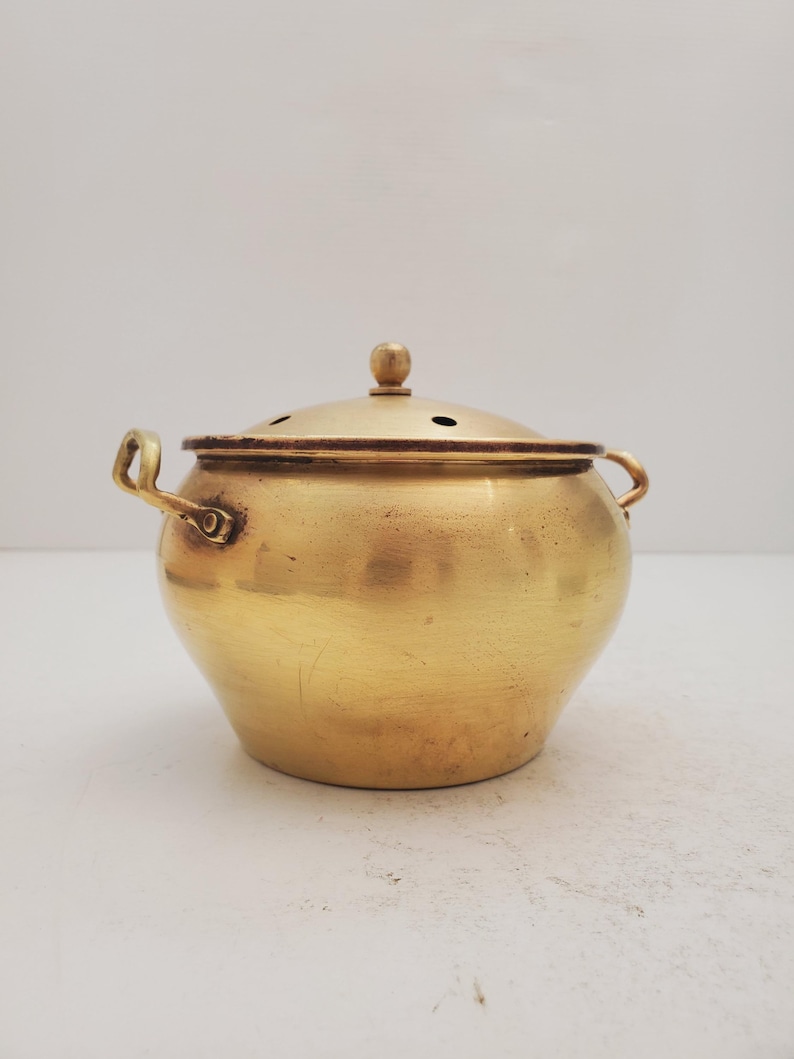 May include: A small, gold-colored brass pot with a lid and two handles. The pot has a round body and a slightly domed lid with small holes.