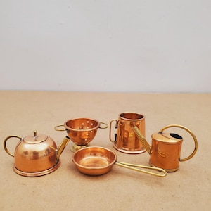May include: A set of miniature copper kitchen utensils, including a teapot, colander, frying pan, mug, and watering can. The items are displayed on a light brown surface, highlighting their shiny, metallic finish and intricate details.