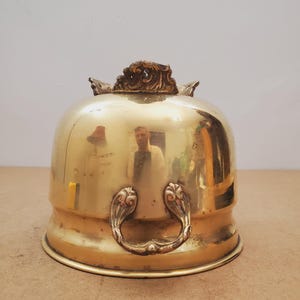May include: A shiny brass cloche with decorative handles and a detailed top. The dome-shaped cover reflects the surroundings. The handles have a floral design. This antique piece is likely used for serving food.