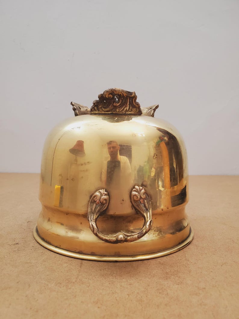 May include: A shiny brass cloche with decorative handles and a detailed top. The dome-shaped cover reflects the surroundings. The handles have a floral design. This antique piece is likely used for serving food.