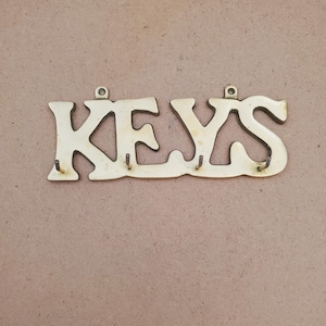May include: A gold-toned metal wall-mounted key holder featuring the word KEYS in a stylized, connected font. The piece includes four small, protruding metal hooks along the bottom edge for hanging items and two circular mounting holes at the top.