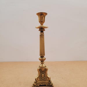 May include: A gold-coloured candlestick with an intricate design. It features a fluted column, a decorative base with a portrait, and a cup-shaped top for a candle. The design is ornate and vintage, suitable for a classic interior.