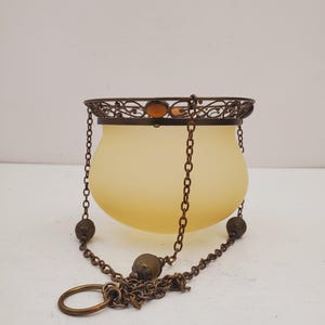 Könnte beinhalten: Eine Vintage-Hängelampe mit einem Schirm aus mattiertem gelbem Glas. Die Lampe hat eine dekorative Metall-Filigran-Oberseite mit einem bernsteinfarbenen Glasakzent. Sie hängt an einer Kette mit dekorativen Metallperlen und einem Ring unten.