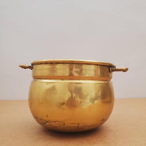 May include: A polished brass pot in a golden hue, featuring two small handles. The pot has a rounded shape and a slightly flared rim. The surface reflects light, creating a warm glow. The pot is placed on a brown surface.