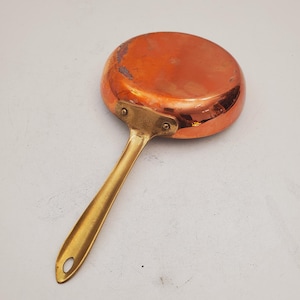 May include: A small copper saucepan with a brass handle. The saucepan has a shiny copper finish and a rounded bottom.