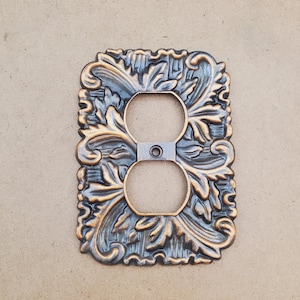 May include: Decorative electrical socket cover with an elaborate, embossed floral pattern. The cover has a two-hole socket design and is finished in bronze and blue-grey hues, offering a vintage appearance. The dimensions are approximately 11 cm x 7 cm.