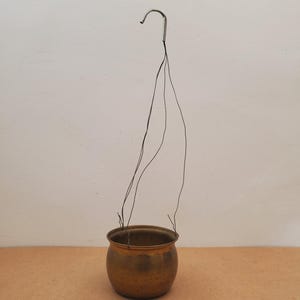 May include: A small, round, antique brass hanging planter with a curved metal hook and thin wire supports. The planter has a smooth, polished finish and is designed to hold a small plant or flowers. The background is a neutral color.