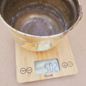 May include: A tarnished brass bowl sits on a wooden digital kitchen scale. The scale displays a weight of 50.2, likely in ounces. The bowl has a rounded shape with a wide opening and a slightly textured surface.