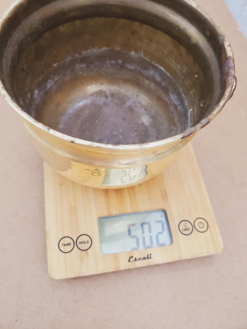 May include: A tarnished brass bowl sits on a wooden digital kitchen scale. The scale displays a weight of 50.2, likely in ounces. The bowl has a rounded shape with a wide opening and a slightly textured surface.