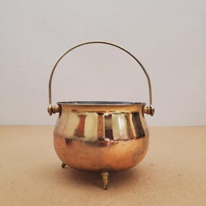 May include: A polished brass cauldron with a rounded body, three small feet, and a thin, arched metal handle attached to the sides. The metallic surface reflects light, showing a warm golden hue and a smooth, curved exterior design.