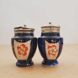 May include: A pair of antique salt and pepper shakers. They are navy blue with silver lids and a floral design in orange and white. The shakers have a small base and are decorated with gold accents.