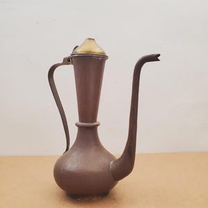 Peut inclure: Une cruche antique de couleur cuivrée avec un long bec courbé et un couvercle de couleur laiton. La cruche a une base bulbeuse et un long col fin. La poignée est une simple pièce de métal incurvée. Le fond est de couleur neutre.