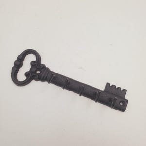 Puede incluir: Gancho de pared en forma de llave de hierro fundido negro con cinco ganchos. La llave tiene un diseño adornado y un borde dentado.