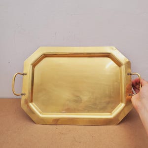 May include: A polished gold-toned brass serving tray with an octagonal shape and a flat base. The tray features two curved metal handles attached to the sides with small rivets, providing a sturdy grip for carrying items on the smooth surface.