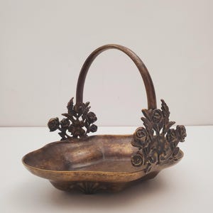 May include: A vintage brass serving basket with a floral design. The basket has a handle and is decorated with roses and leaves.