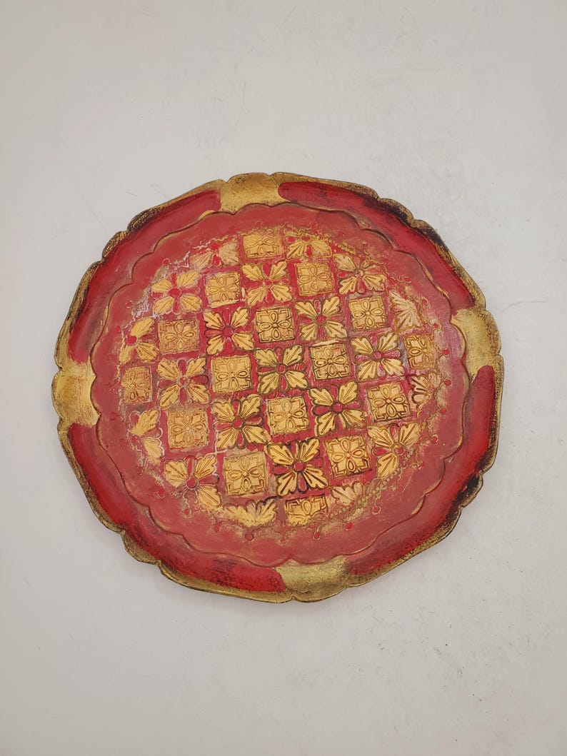K&ouml;nnte beinhalten: Ein rundes, rotes Holztablett mit einem goldfarbenen Design aus sich wiederholenden Quadraten und Blumen. Das Tablett hat einen gewellten Rand.