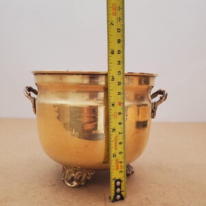 May include: A gold-coloured metal bowl with ornate handles and decorative feet. The bowl is approximately 18 cm tall, as measured by a yellow tape measure. The bowl has a rounded shape and a polished finish.