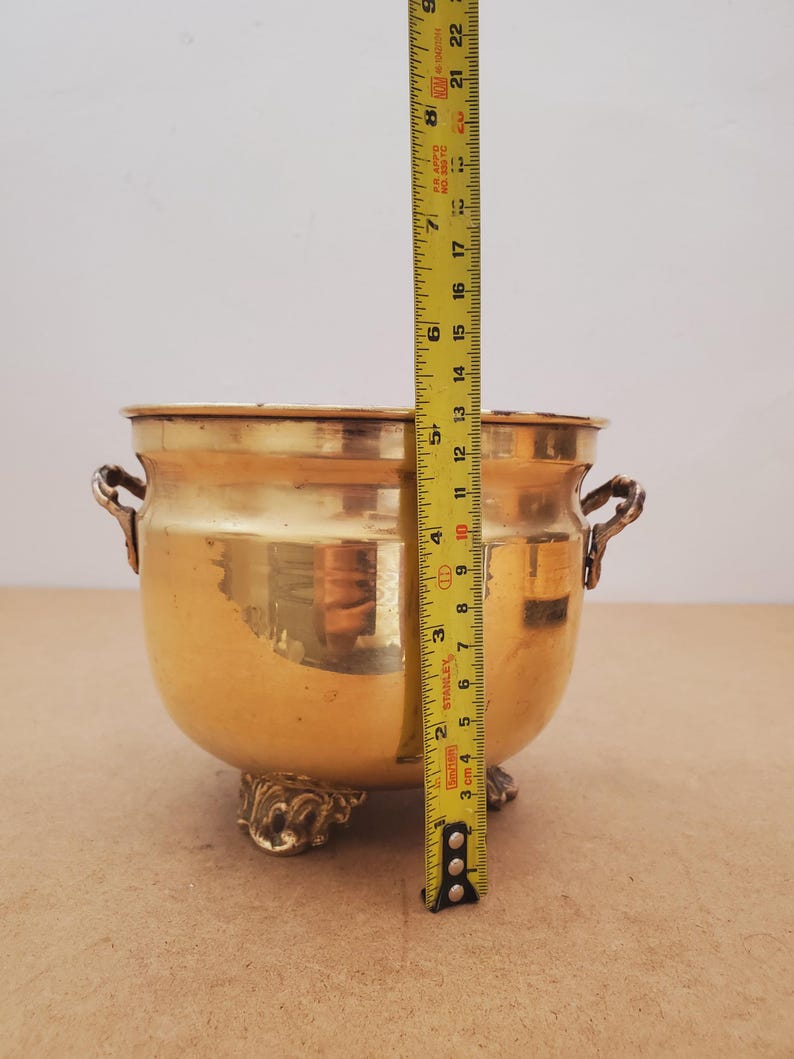 May include: A gold-coloured metal bowl with ornate handles and decorative feet. The bowl is approximately 18 cm tall, as measured by a yellow tape measure. The bowl has a rounded shape and a polished finish.