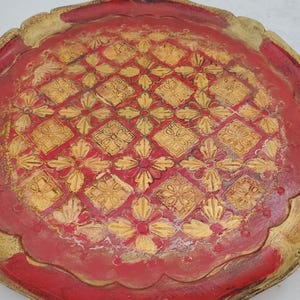 K&ouml;nnte beinhalten: Ein rundes, rotes Holztablett mit einem goldfarbenen Rand und einem dekorativen Muster aus goldenen Blumen und Quadraten.