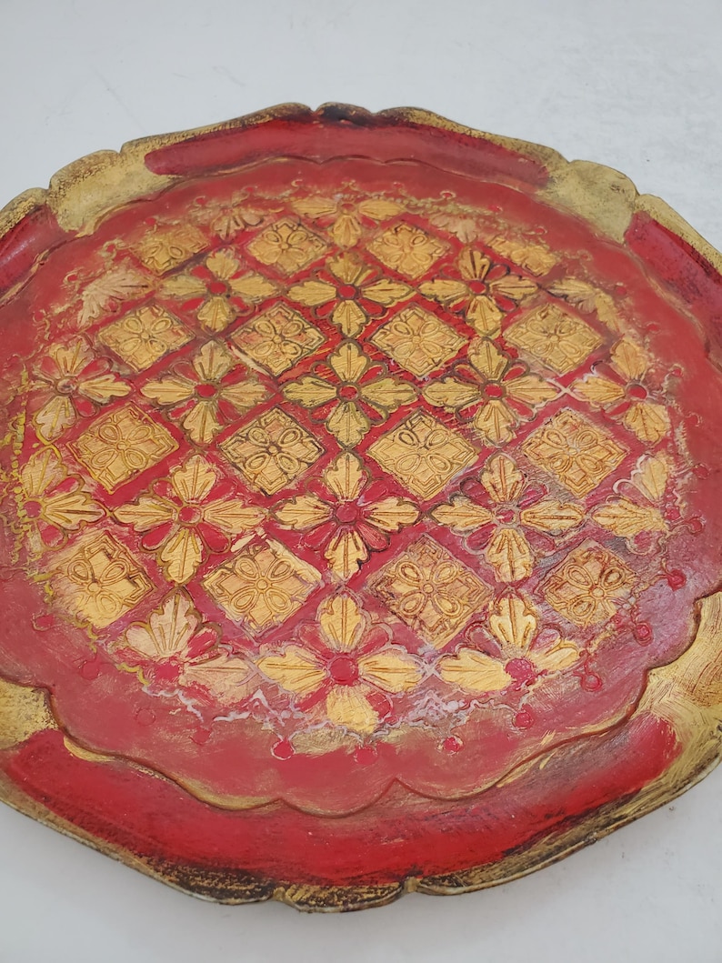 K&ouml;nnte beinhalten: Ein rundes, rotes Holztablett mit einem goldfarbenen Rand und einem dekorativen Muster aus goldenen Blumen und Quadraten.