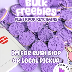 Op de afbeelding: Een verzameling ronde, paarse, koekvormige sleutelhangers met reliëfmotieven, waaronder het BTS-logo, vingerhartjes en lichtstaven. Elke sleutelhanger heeft een witte tussenlaag en een paarse kogelketting om hem aan op te hangen.