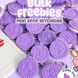 May include: A collection of purple cookie-shaped keychains featuring embossed K-pop symbols and logos. The text BULK FREEBIES MINI KPOP KEYCHAINS is overlaid in white. Each keychain has a textured edge and a small ball chain attachment.