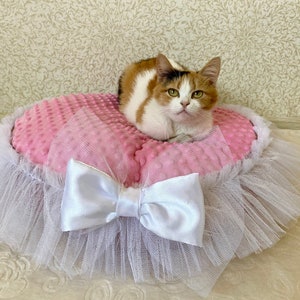 May include: A pink and white cat bed with a white tulle skirt and a white satin bow. A calico cat is sitting on the bed.