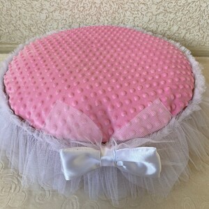 May include: A pink and white pet bed with a white tulle ruffle and a white satin bow. The bed is made of soft, plush fabric with a dotted pattern.