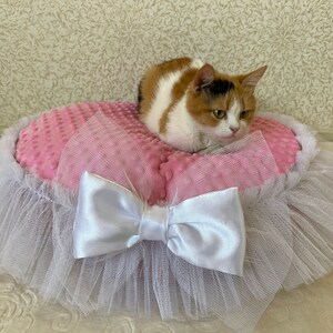 May include: A pink and white cat bed with a white tulle skirt and a white satin bow. A calico cat is sitting on the bed.