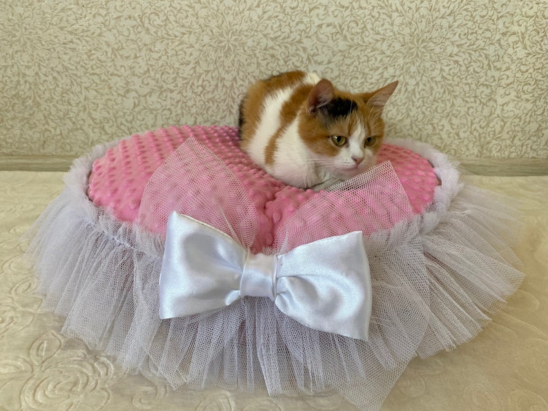 May include: A pink and white cat bed with a white tulle skirt and a white satin bow. A calico cat is sitting on the bed.