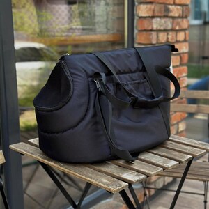 Puede incluir: Transport&iacute;n acolchado negro para mascotas con una ventana de malla y correas ajustables. El transport&iacute;n est&aacute; sobre una mesa de madera.