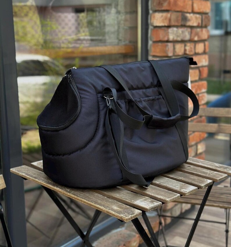 Puede incluir: Transport&iacute;n acolchado negro para mascotas con una ventana de malla y correas ajustables. El transport&iacute;n est&aacute; sobre una mesa de madera.