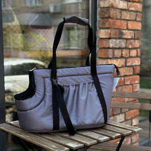 Puede incluir: Un transport&iacute;n para mascotas gris con correas negras y un interior acolchado. El transport&iacute;n tiene un bolsillo con cremallera en la parte delantera y un asa en la parte superior.