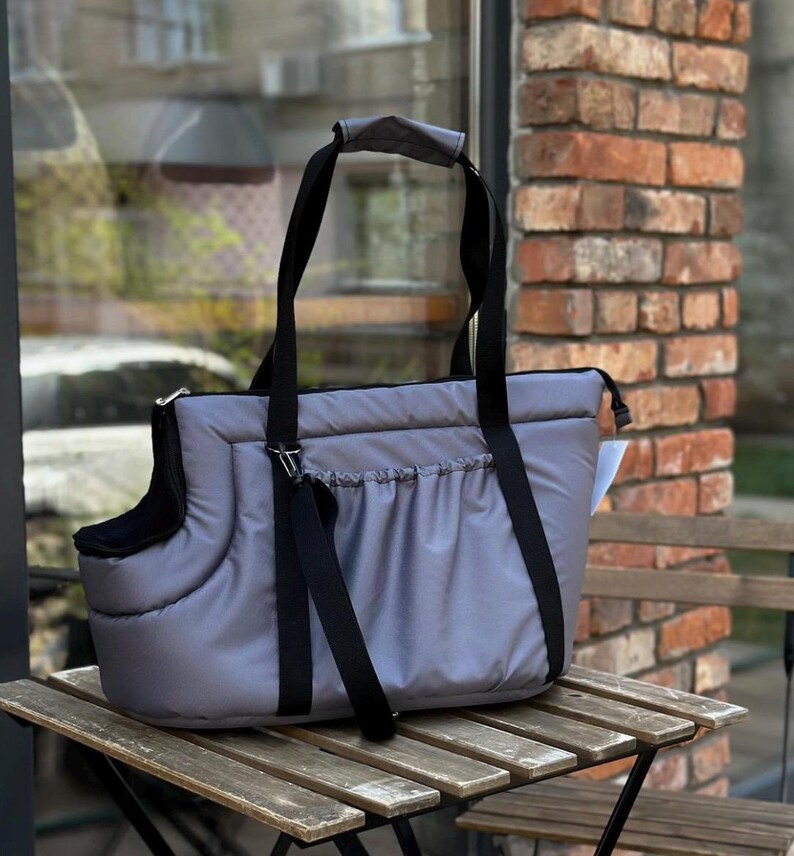 Puede incluir: Un transport&iacute;n para mascotas gris con correas negras y un interior acolchado. El transport&iacute;n tiene un bolsillo con cremallera en la parte delantera y un asa en la parte superior.