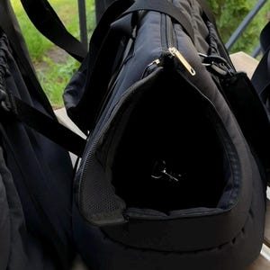 Puede incluir: Bolsa de transporte para mascotas negra con una abertura con cremallera y un panel de malla. La bolsa tiene un asa y una correa para el hombro.