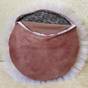 May include: A round, blush pink velvet cushion with a white tulle ruffle around the edge. The cushion has a zipper closure and is filled with a gray lace fabric.