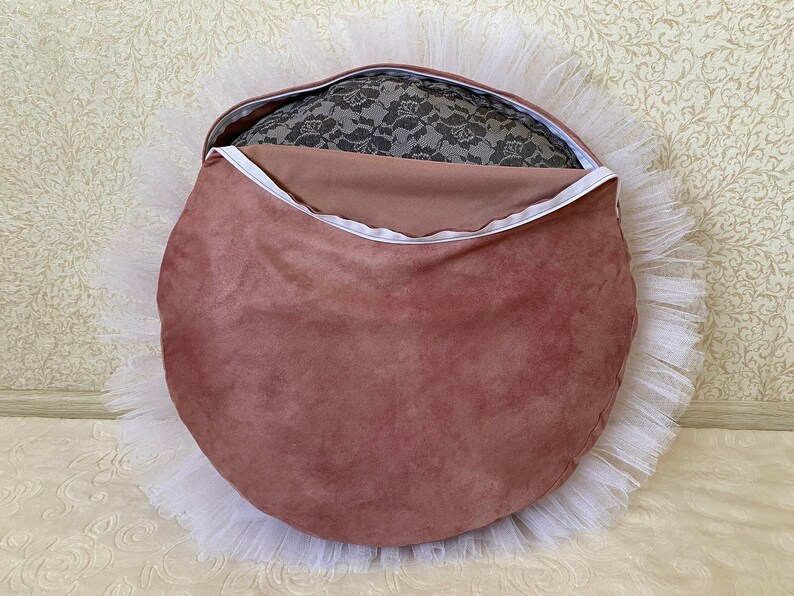 May include: A round, blush pink velvet cushion with a white tulle ruffle around the edge. The cushion has a zipper closure and is filled with a gray lace fabric.