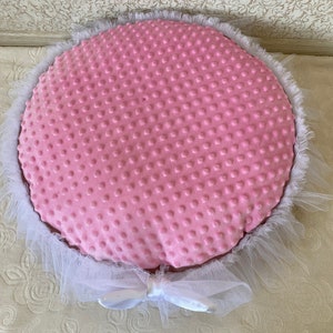 May include: A pink round pet bed with a white tulle ruffle around the edge. The bed has a soft, textured fabric with a dotted pattern.