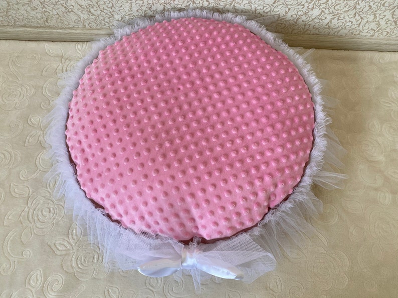 May include: A pink round pet bed with a white tulle ruffle around the edge. The bed has a soft, textured fabric with a dotted pattern.