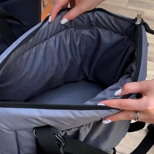 Puede incluir: Un transport&iacute;n gris para mascotas con una cremallera negra y un asa negra. El transport&iacute;n est&aacute; abierto y el interior est&aacute; forrado con un tejido gris suave.