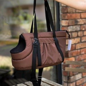 Puede incluir: Un transport&iacute;n para mascotas de color marr&oacute;n con una correa negra y una ventana de malla negra. El transport&iacute;n tiene un bolsillo en la parte delantera y una abertura con cremallera en la parte superior.