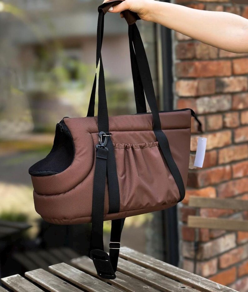 Puede incluir: Un transport&iacute;n para mascotas de color marr&oacute;n con una correa negra y una ventana de malla negra. El transport&iacute;n tiene un bolsillo en la parte delantera y una abertura con cremallera en la parte superior.