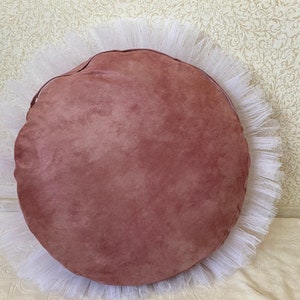 May include: A round, blush pink suede cushion with a white tulle ruffle around the edge.