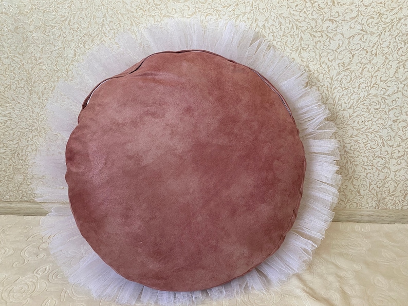 May include: A round, blush pink suede cushion with a white tulle ruffle around the edge.