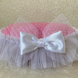May include: A pink and white round pillow with a white satin bow and a white tulle ruffle around the edge.