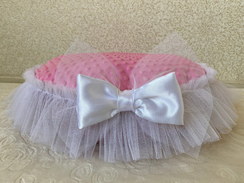 May include: A pink and white round pillow with a white satin bow and a white tulle ruffle around the edge.